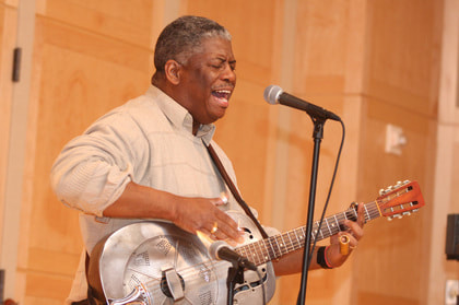 a black man with short greying hair singing into a microphone and playing a silver steel guitar
