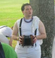 a white person with short dark hair, wearing white pants and a white shirt with black suspenders. Zie is singing and playing a concertina