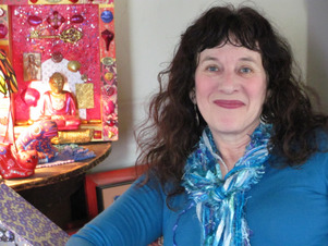 a woman with white skin and dark hair, sitting next to a bright pink piece of art with a buddha statue in it, looking into the camera with a smile