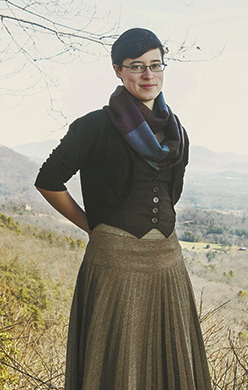 a young person with white skin, dark hair, and glasses, wearing a black vest and a grey skirt and smiling slightly into the camera