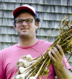 a white man with glasses and a baseball cap, holding a bundle of dried garlic heads and looking into the camera and smiling
