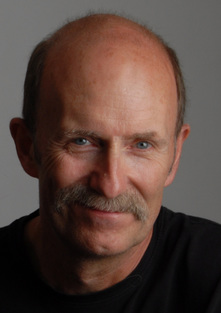 a white balding man with a grey mustache and grey eyes, looking directly into the camera with a slight smile