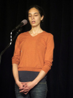 a white woman with dark hair wearing an orange sweater and jeans singing into a microphone