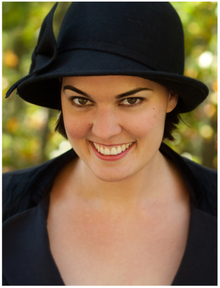 a white woman wearing a dark hat and a scoop-neck dark shirt, looking slightly up into the camera with a wide smile