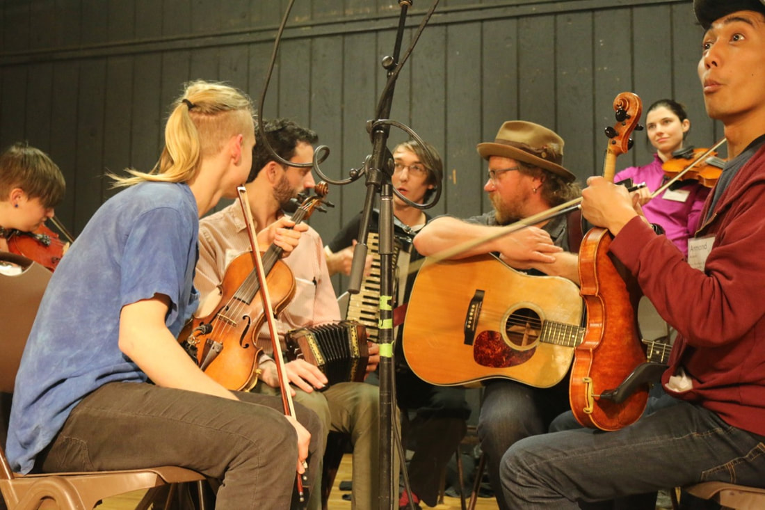 a group of people sit in a circle holding instruments, including fiddles, a guitar, and an accordion