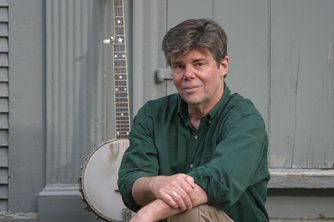 a white man with greying short hair, wearing a green shirt and sitting with his arms crossed. he is looking into the camera and a banjo is visible behind him
