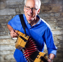 a white man with glasses and white hair, wearing a bright blue shirt and holding an accordian, looking into the camera with a wide smile