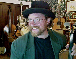 a white man with glasses and a blonde beard, wearing a green jacket and a dark hat and holding a fiddle