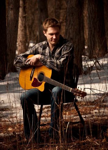 a young white man sitting on a chair outside in the snowy woods holding a guitar and looking to the side