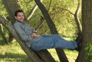 a white man with dark hair and a beard, sitting stretched out in a spreading tree