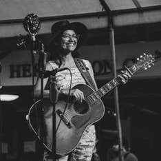 a black and white image of a woman with dark tan skin, dark hair, and glasses. She is wearing a wide-brim hat and overalls, standing on stage smiling and holding a large acoustic guitar