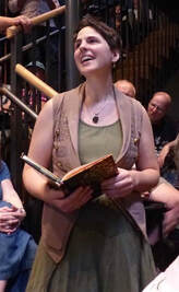 a white woman with short brown hair stands and sings while holding a book