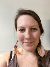 a white woman with long brown hair and large feather earrings, looking directly into the camera with a small smile