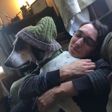 a young white person with glasses and dark hair, hugging a grey dog with a green knit hat on its head