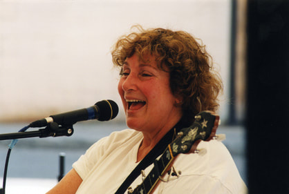 a white woman with curly grown hair singing happily into a microphone and holding a banjo