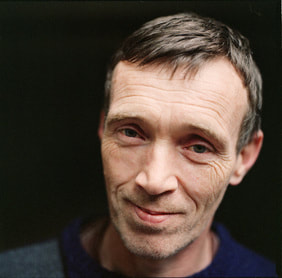 a white man with short brown hair and a wrinkled face, looking into the camera and smiling gently